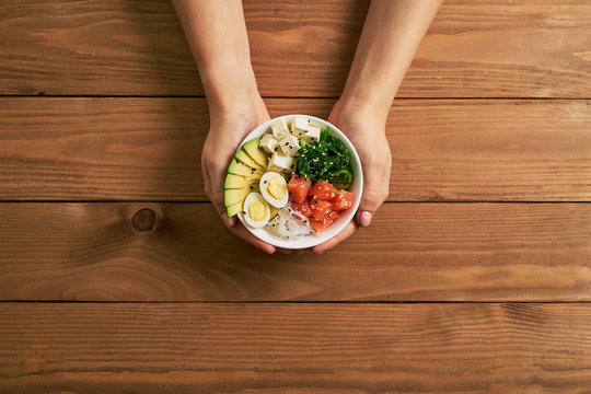 Female Hands Holding Poke Bowl With Salmon, Avocado Rice, Chuka Salad, Sweet Onions, Quail Eggs Sprinkled With White And Black Sesame Soy Sauce Isolated On Wooden Background Top View