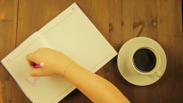 The girl writes in the diary the love word with a pink marker and drinks coffee.