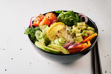 poke bowl with salmon, avocado, cucumber, arugula, broccoli, rice, carrot and sweet onions with chopsticks isolated over white background. side view
