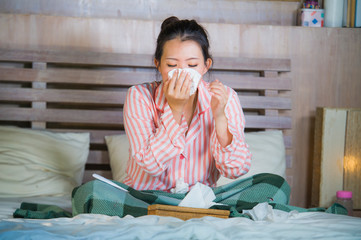 sweet Asian American woman in pajamas sick at home bedroom suffering cold and flu taking temperature with thermometer in bed blowing her nose coughing and sneezing