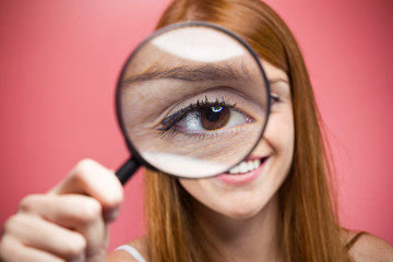 Cheerful pretty girl looking through magnifying glass isolated over pink background