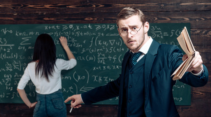 Genius man in glasses with book point at woman solving equation on chalkboard. Genius teacher with student in classroom