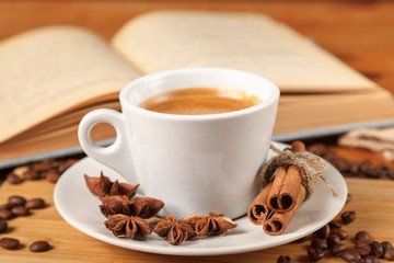 Coffee break a Cup of hot coffee and a book on a wooden table. A white Cup of black coffee surrounded by a small amount of roasted coffee beans, cinnamon, anise and an open book