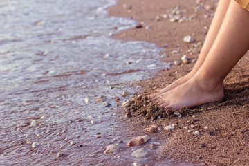 female feet on wet sand sea shoreline surface near water of waves in evening sunset time with soft orange color filter, summer vacation and rest concept
