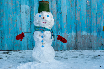 White Snowman dressed in scarf, tin bucket with blue eyes and carrot nose against background of blue wooden fence