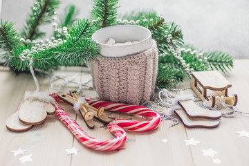 White cup for cocoa in beige knitted clothes. Santa Claus sleigh, mittens, hat, sweater and socks on a white wooden background with christmas tree and red caramel candy