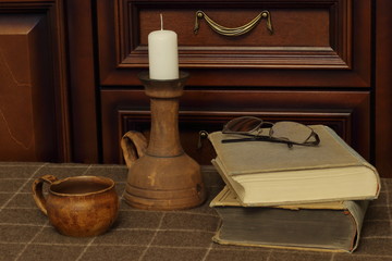 Rare candlestick with candle, old clay cup, antique books and glasses on woolen plaid against background of wooden secretary.