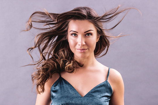 Fashion portrait of gorgeous young woman standing against gray background with hair in motion. Beautiful fashionable girl in party dress looking to camera and smiling while isolated. Waving hair,Vogue