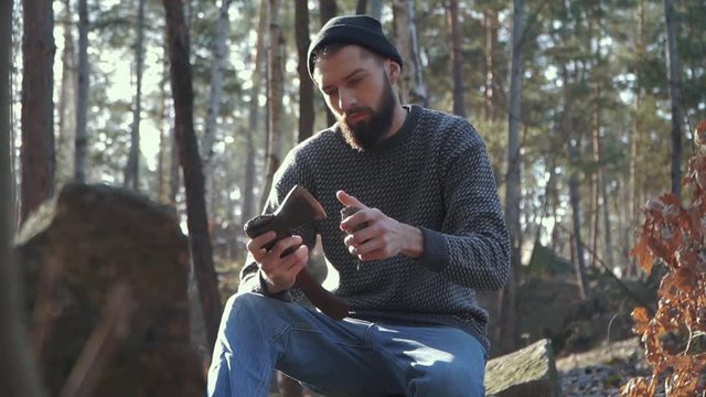 Young bearded guy sharps an axe in the forest. Brutal man sitting with an ax outdoors.