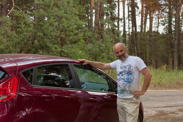 A man of mature age in the forest by the road put his hand on the body of the car