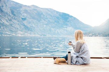 Girl on wooden pier by winter sea, mountains. Cozy picnic with hot beverages, tea, coffee or cocoa in thermos and mug, warm plaid, opened book. Concept of enjoying nature, relaxation, reading on beach