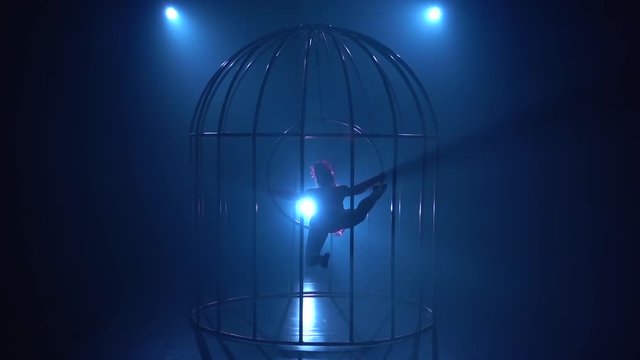 Silhouette of a girl spinning on a hoop on stage in a cages. Blue smoke background. Silhouette. Slow motion