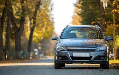 Front view of gray shiny empty car parked in quiet area on asphalt road at lamppost on blurred green and yellow trees bokeh background on bright sunny day. Transportation and parking concept.