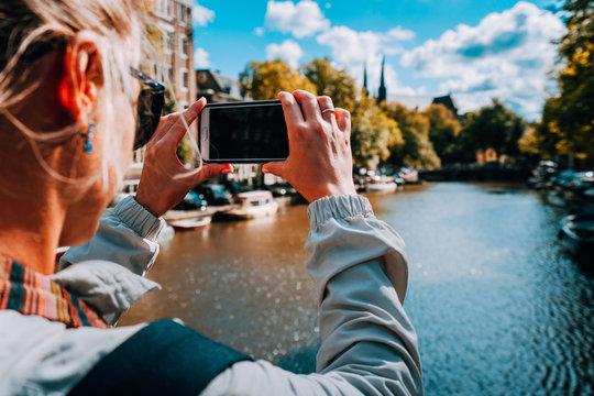 Rear view of female tourist taking photo of canal in Amsterdam on the mobile phone on sunny autumn day. Warm gold afternoon sunlight. Travel in Europe