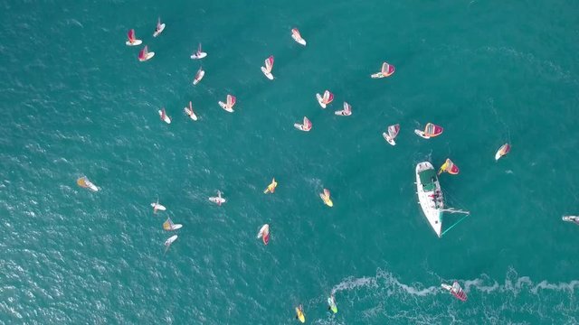Small white yacht surrounded by a large group of Wind surfers at The Mediterranean Sea - Top down aerial footage.