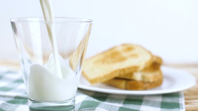 Milk pouring in glass with toast bread on white plate and green tablecloth on wooden kitchen surface. Slow motion footage.