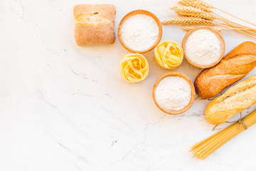 Farinaceous food. Fresh bread and raw pasta near flour in bowl and wheat ears on white stone background top view space for text