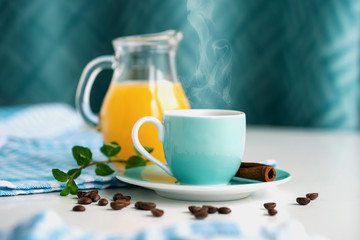 Morning coffee in a blue cup beside cinnamon and scattered coffee beans, in the background a jug with orange juice.