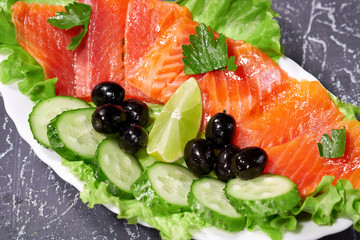 Sliced salmon pieces on a white plate with olives and lemon, on a grey background. Selective focus.