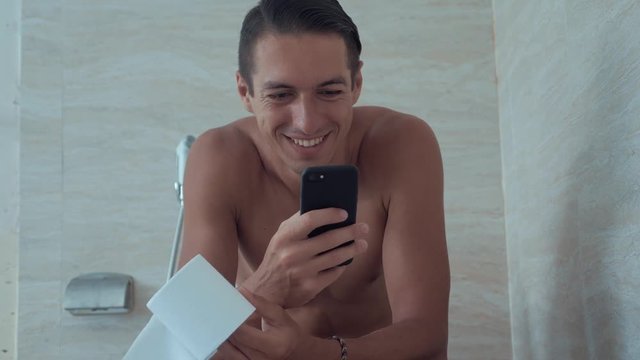 Young man uses smart phone sitting on toilet at home. Man sitting in toilet with phone.