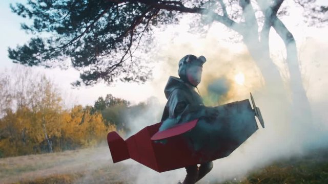 Camera follows little happy aviator boy running in forest wearing cardboard plane costume with color smoke slow motion.