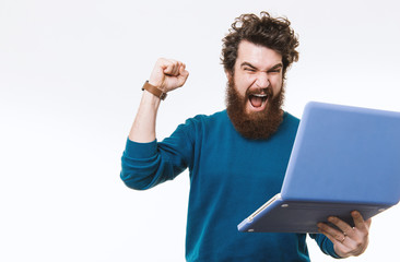 Amazed bearded hipster man clebrating success and looking at laptop over white background, studio...