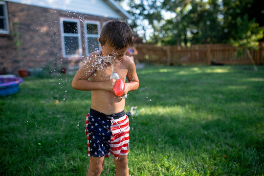 Shirtless boy playing with water bombs while standing on grassy field at backyard