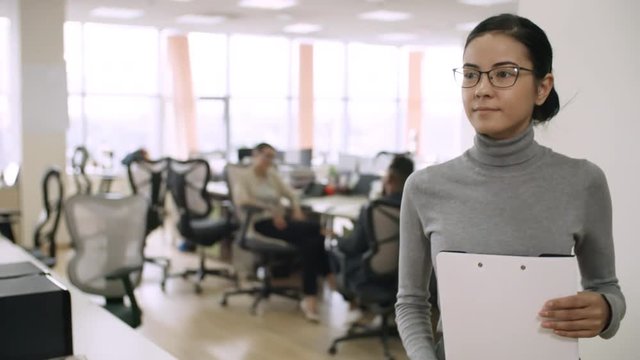 Medium shot of young mixed race woman in glasses carrying clipboard with documents through modern office, slow motion
