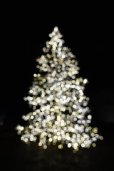 Silhouette of blurry Christmas tree illuminated and decorated with golden, silver and white fairy string lights in dark night creating beautiful bokeh effect with glowing and shiny metallic circles