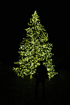 Blurry Silhouette Of Man Standing In Front Of An Looking At Illuminated Christmas Tree With Green Fairy String Lights Creating Beautiful Out Of Focus Bokeh In Dark Night