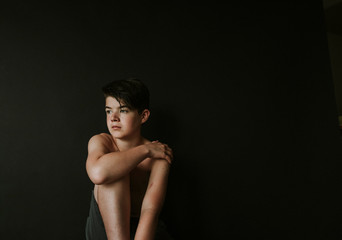 Thoughtful shirtless boy looking away while sitting against black background