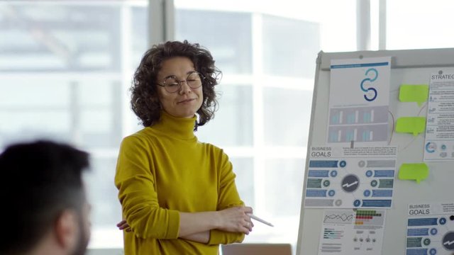 Tilt down of young caucasian businesswoman in eyeglasses explaining scheme of business goals and strategy on flipchart while giving presentation to colleagues during office meeting