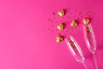 Champagne glasses and heart shaped candies in golden foil on color background, top view