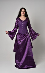 full length portrait of beautiful girl with long black hair,   wearing purple fantasy medieval gown. standing pose on grey studio background.