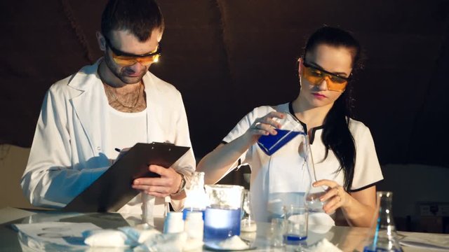 A woman has dressed goggles. Laborant is examining the reaction of a substance in a test tube while stirring. Man is writing the results of experiments in the room near the table