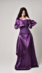 full length portrait of beautiful girl with long black hair,   wearing purple fantasy medieval gown. standing pose on grey studio background.