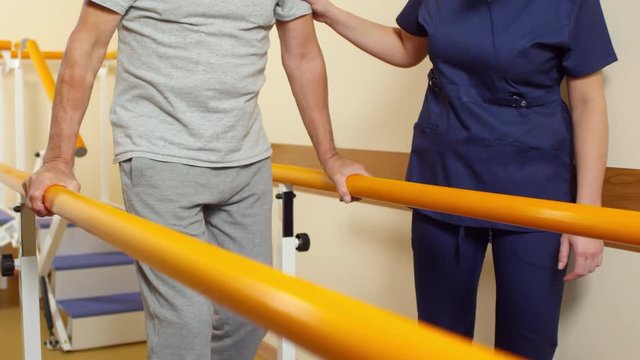 Tilt down shot of elderly man walking between handrails and supporting his weight while learning to walk with help of female physiotherapist