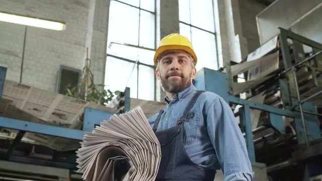 Zoom in of bearded male technician in overalls and hardhat walking in printing plant with stack of newspapers, looking at camera and smiling