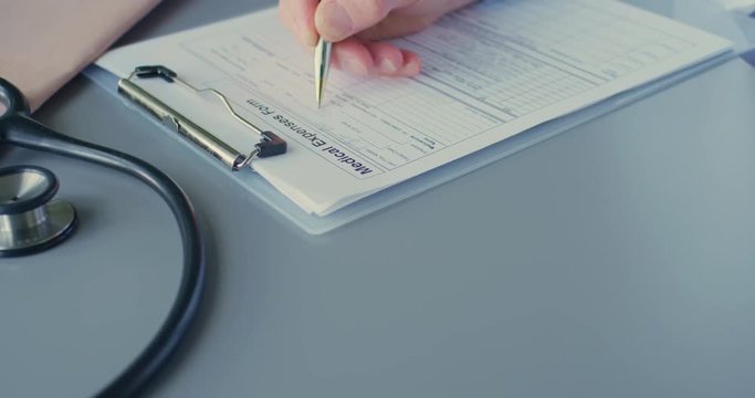 Doctor Filling Medical Expenses Form On Clipboard At Desk 4K