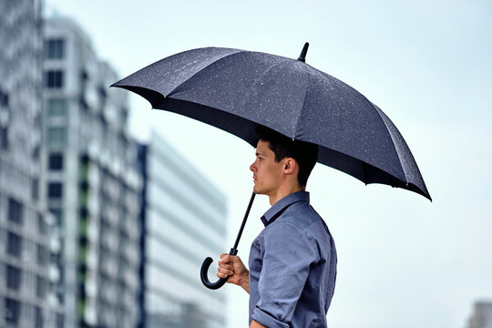 Side view of young businessman with umbrella standing against buildings and sky in city during rainfall
