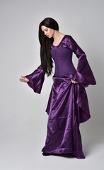 Naklejka premium full length portrait of beautiful girl with long black hair, wearing purple fantasy medieval gown. standing pose on grey studio background.