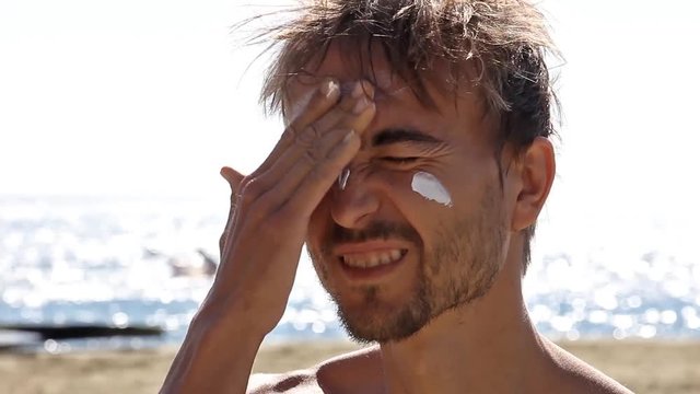 Handsome bearded man applies sunscreen on his face. Sea, beach and windsurfers in the background. 