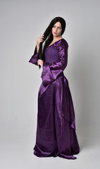 full length portrait of beautiful girl with long black hair,   wearing purple fantasy medieval gown. standing pose on grey studio background.