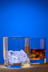 Two glasses on the table, first with pieces of ice and second with drink on a blue background