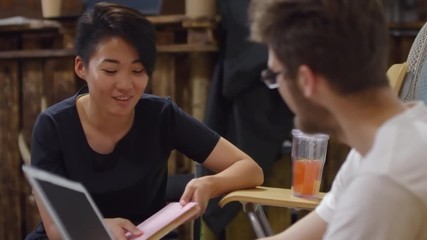 Tilt up shot of young Asian woman holding book and talking to man seen from his back during break at business or IT seminar
