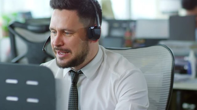 Medium shot of bearded Caucasian man wearing headset and using desktop computer when helping client online at customer support