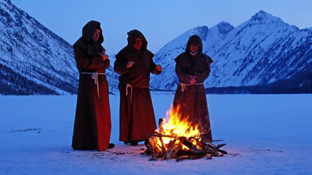 Group of monks in brown cloaks of the order of St. Francis are warming themselves by the fire