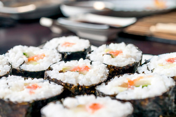 Chumaki Sushi Rolls with Tuna, Salmon, Rice, Cucumber, Avocado And Nori Seaweed on a Plate with Soy Sauce Dipping Dish and Kitchenware Blurred in Background.