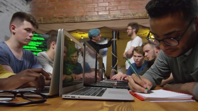 Medium shot of young men of different ethnicities sitting at desk and learning to write codes in programming class