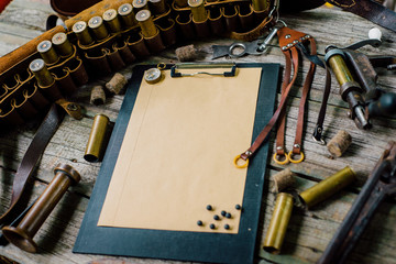 Clipboard with paper and pellets on it on old wooden background. Hunting equipment on vintage desk. Hunting belt with cartridges and tools.